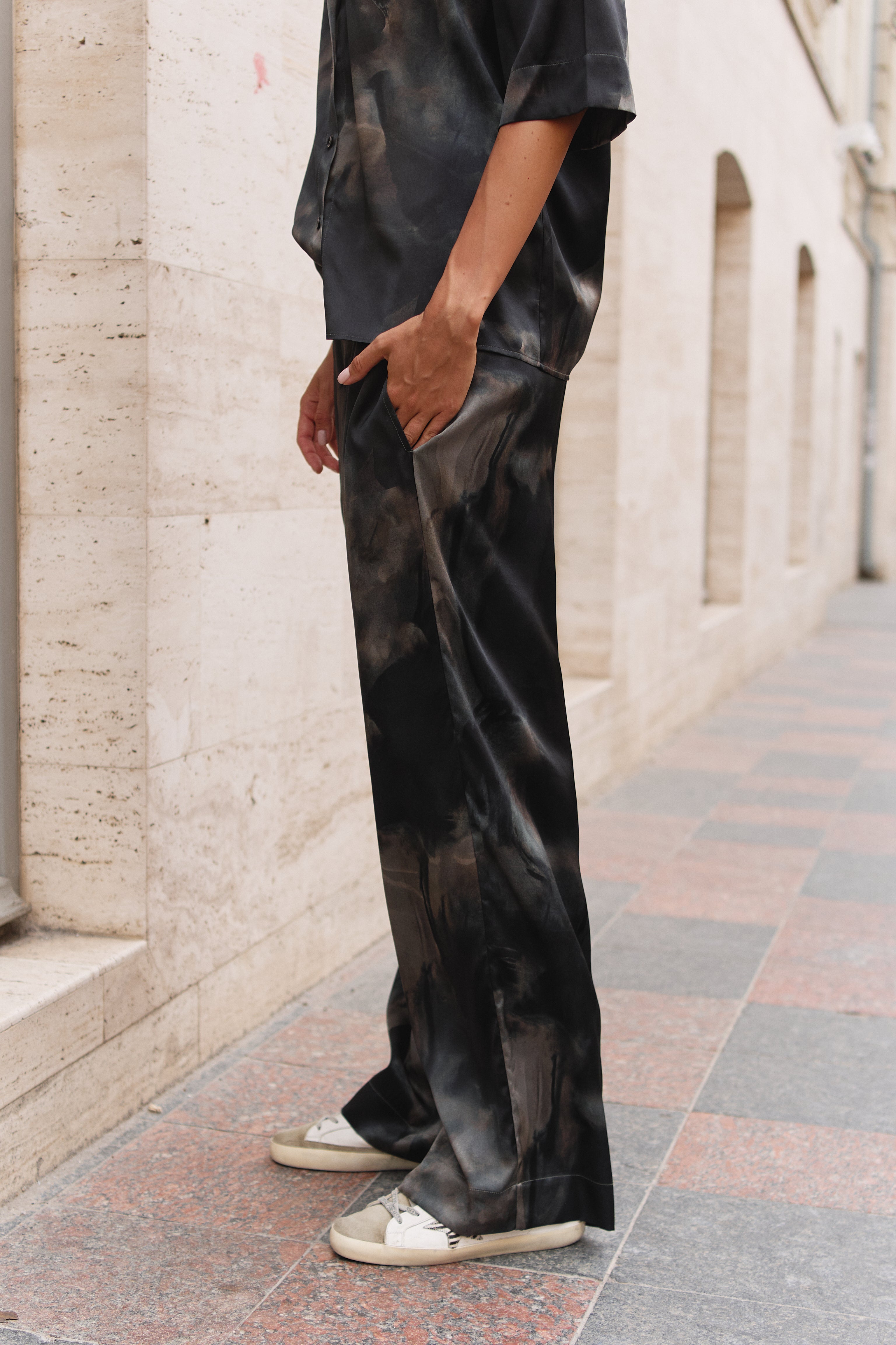 A person standing outdoors on a paved walkway beside a light stone building, wearing loose black satin-like pants with a subtle tonal marbled pattern and a dark short-sleeve top.