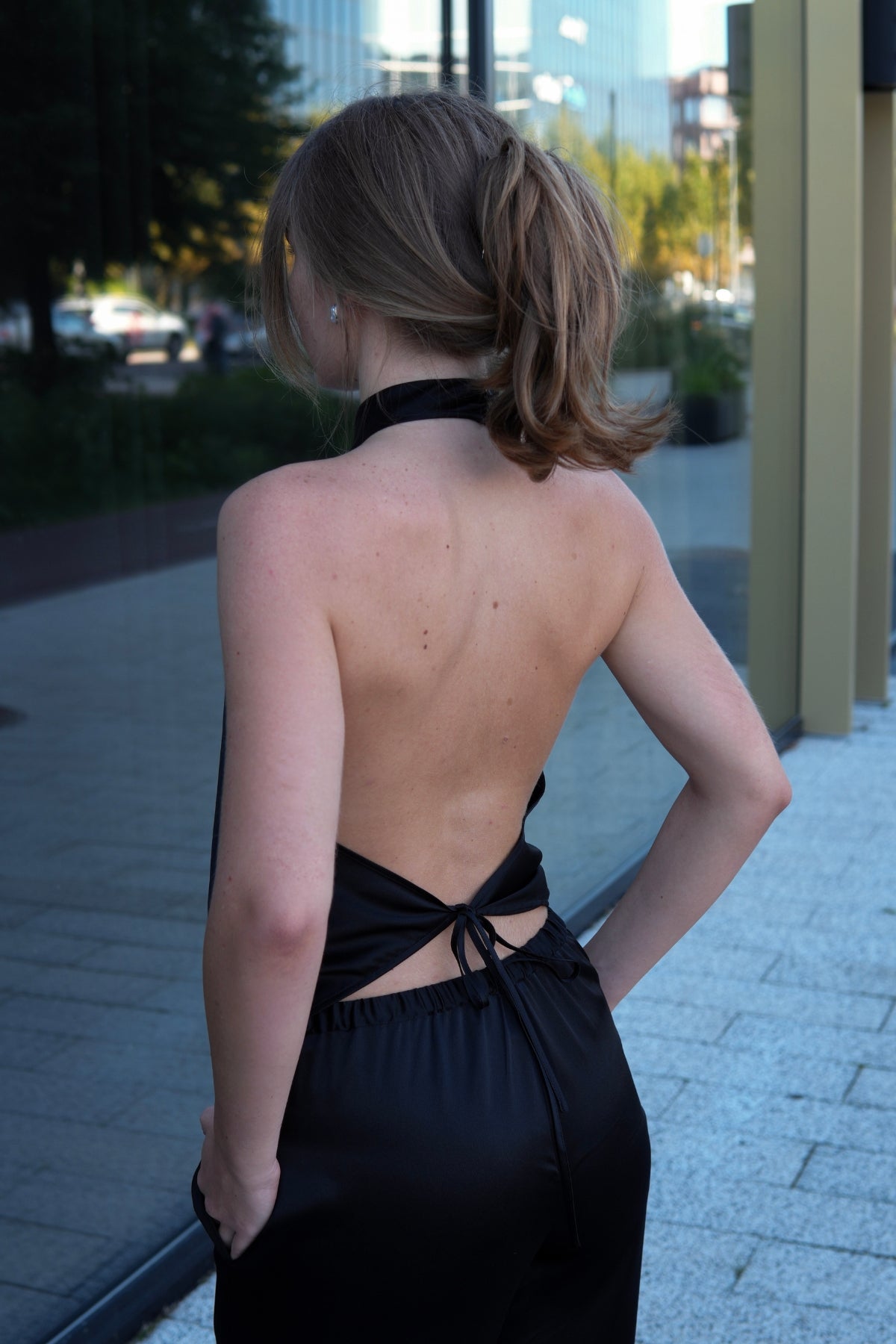 Вlack silk halter-neck top with an open back and tie detail, standing outdoors on a paved walkway.