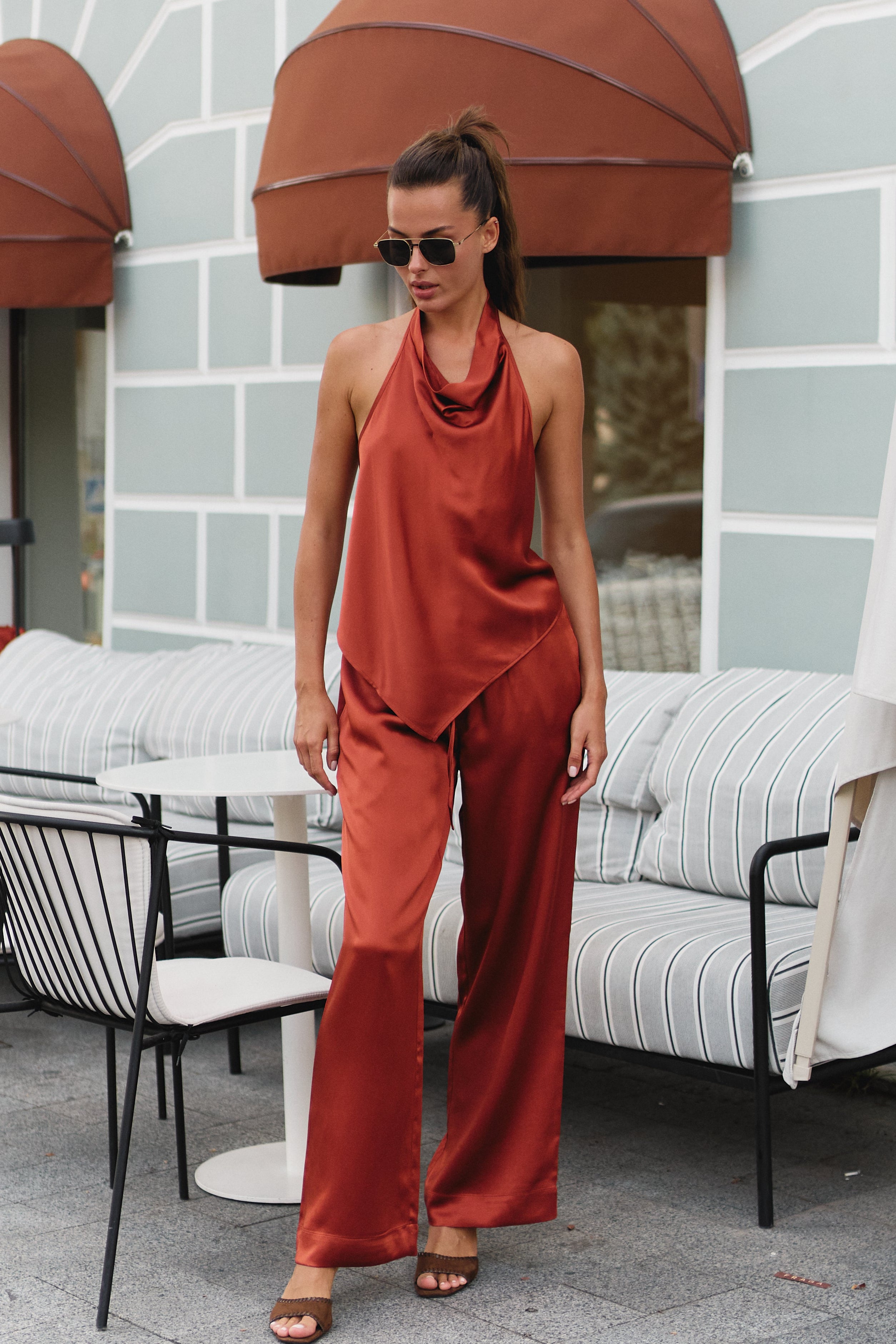 Silk rust-orange halter top Nokaya and matching wide-leg pants stand on an outdoor patio in front of a light grey wall with brown awnings and striped seating.