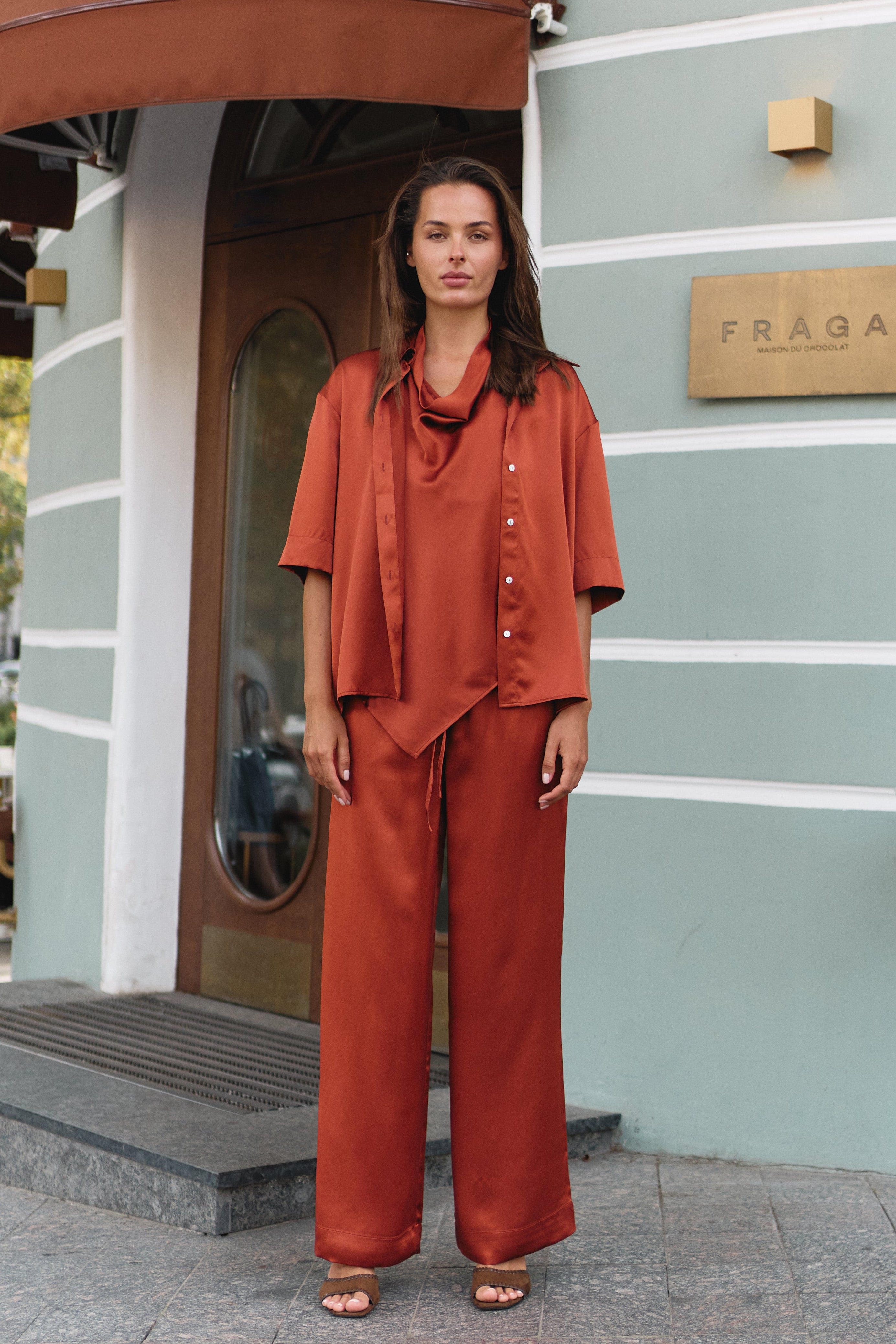Silk matching rust-orange set Nokaya consisting of a short-sleeve button-up shirt and wide-leg pants, standing in front of a modern building with a light green facade.