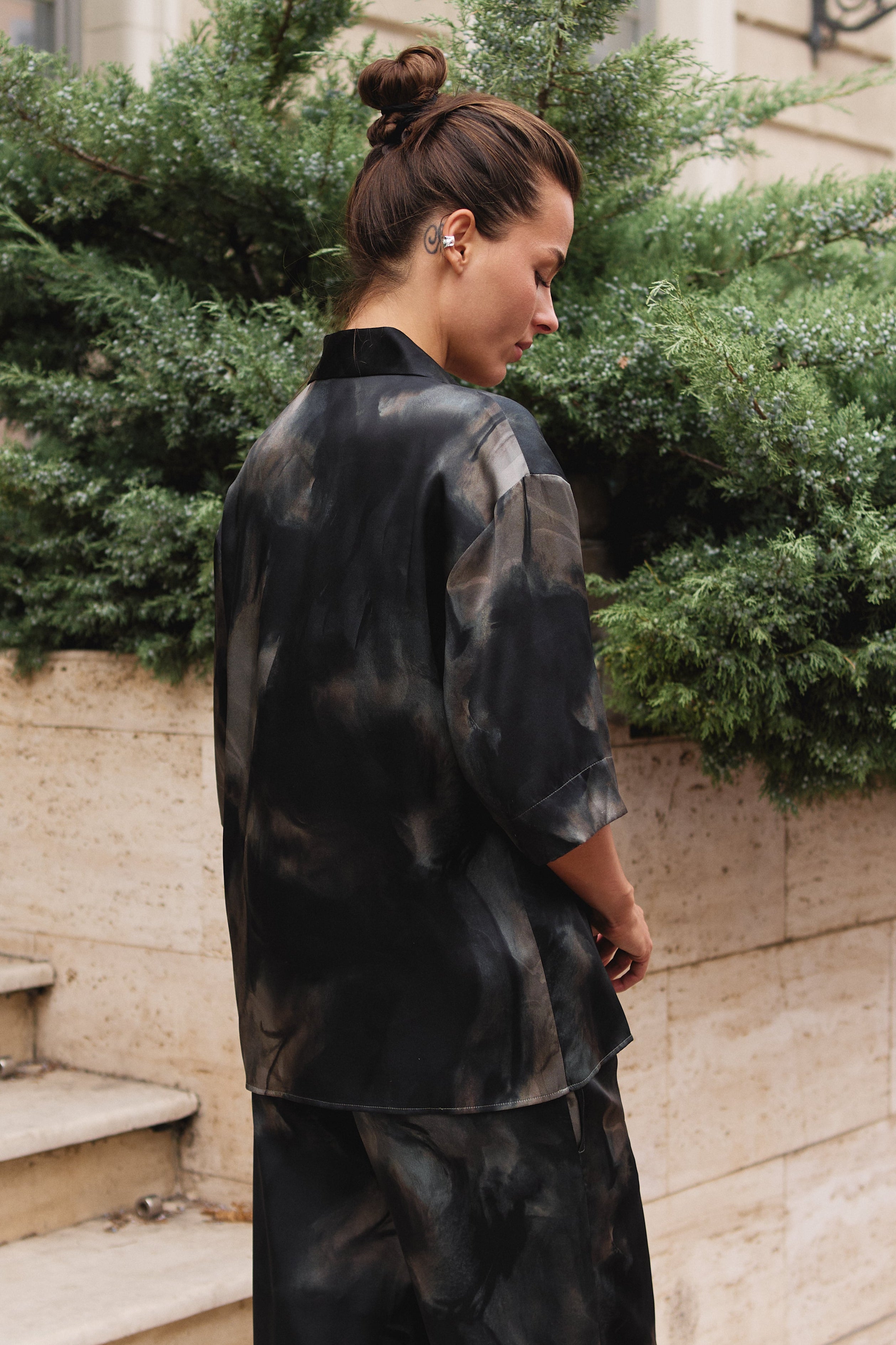 black and grey tie-dye matching silk set Nokaya, photographed from behind as she stands outdoors next to a beige stone wall and green plants.