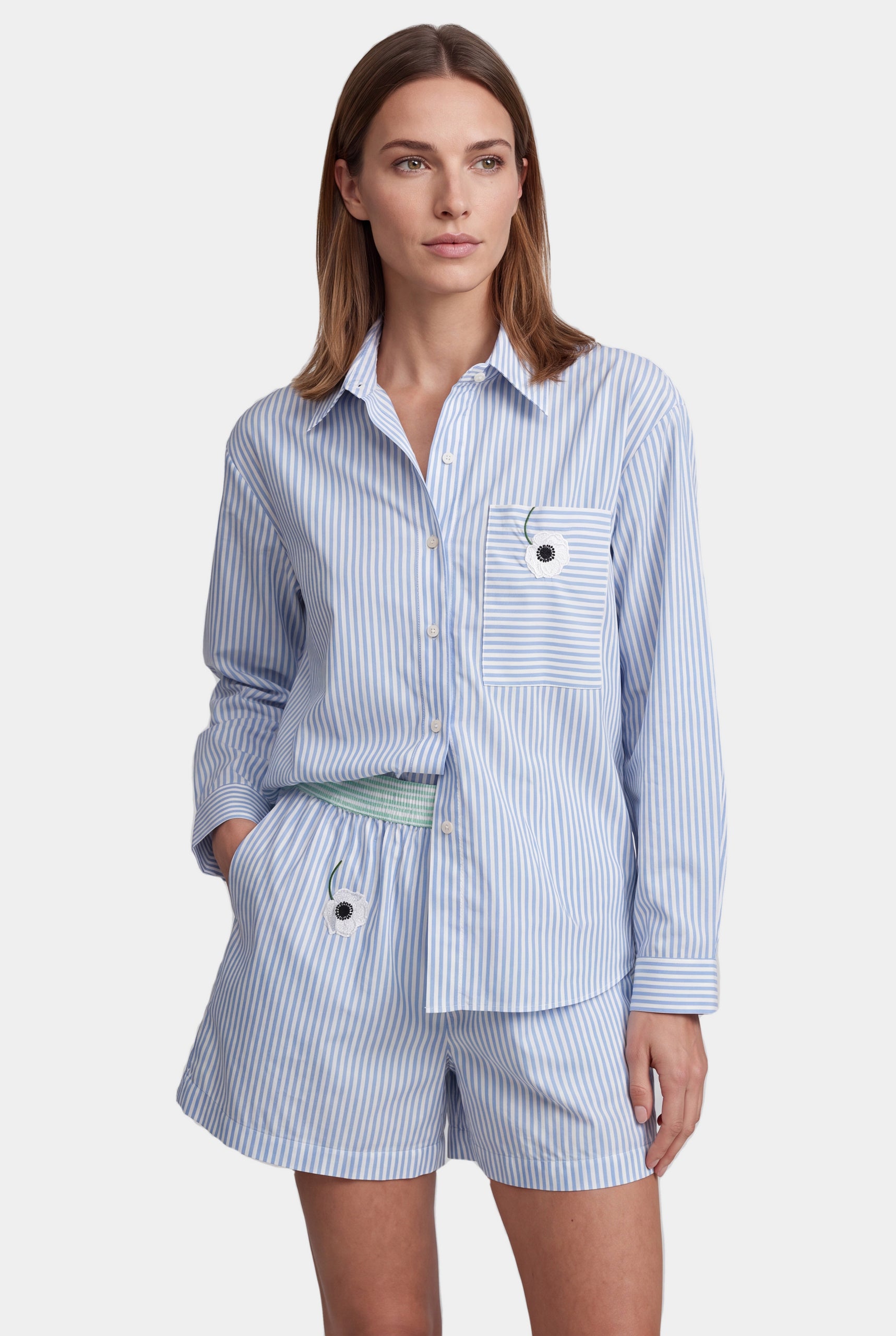 A woman modeling a light blue and white striped matching lounge set consisting of a long-sleeve button-up shirt and shorts, with a light green patterned waist tie belt, against a light gray background.