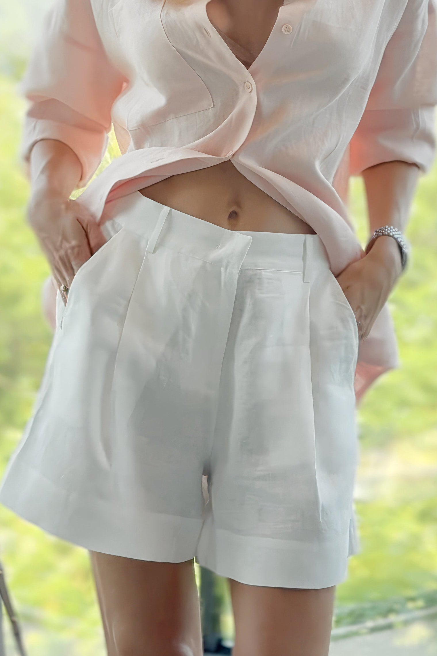 White linen shorts from the Linen Spirit capsule Nokaya, standing outdoors near a window with green foliage in the background. She is wearing sunglasses and has one hand in her pocket.