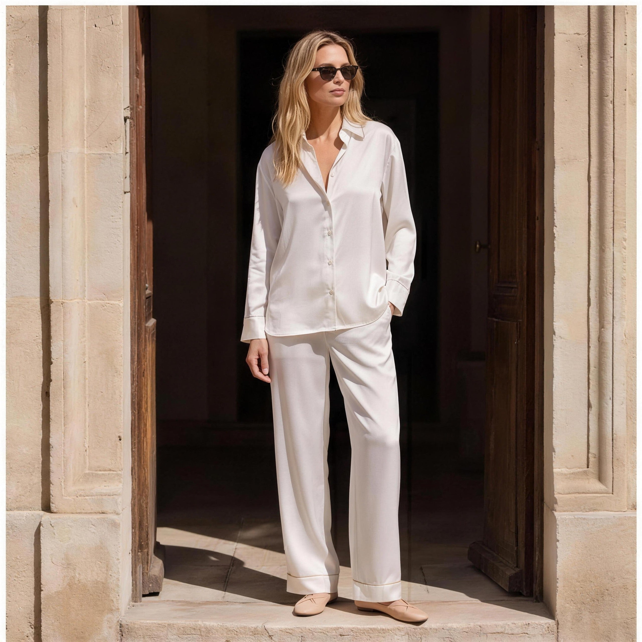 Woman wearing ivory silk pyjama set in a sunlit stone doorway, NOKAYA