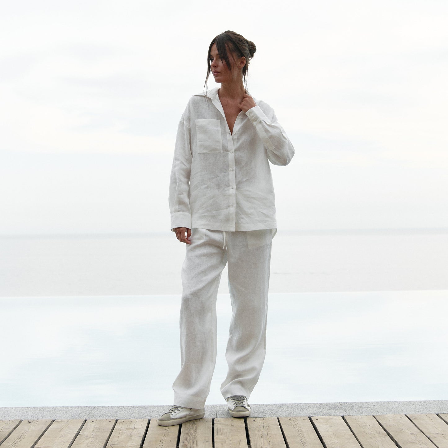A woman wearing a white linen outfit is standing on a wooden deck with a white background. Linen Spirit collection by Nokaya.