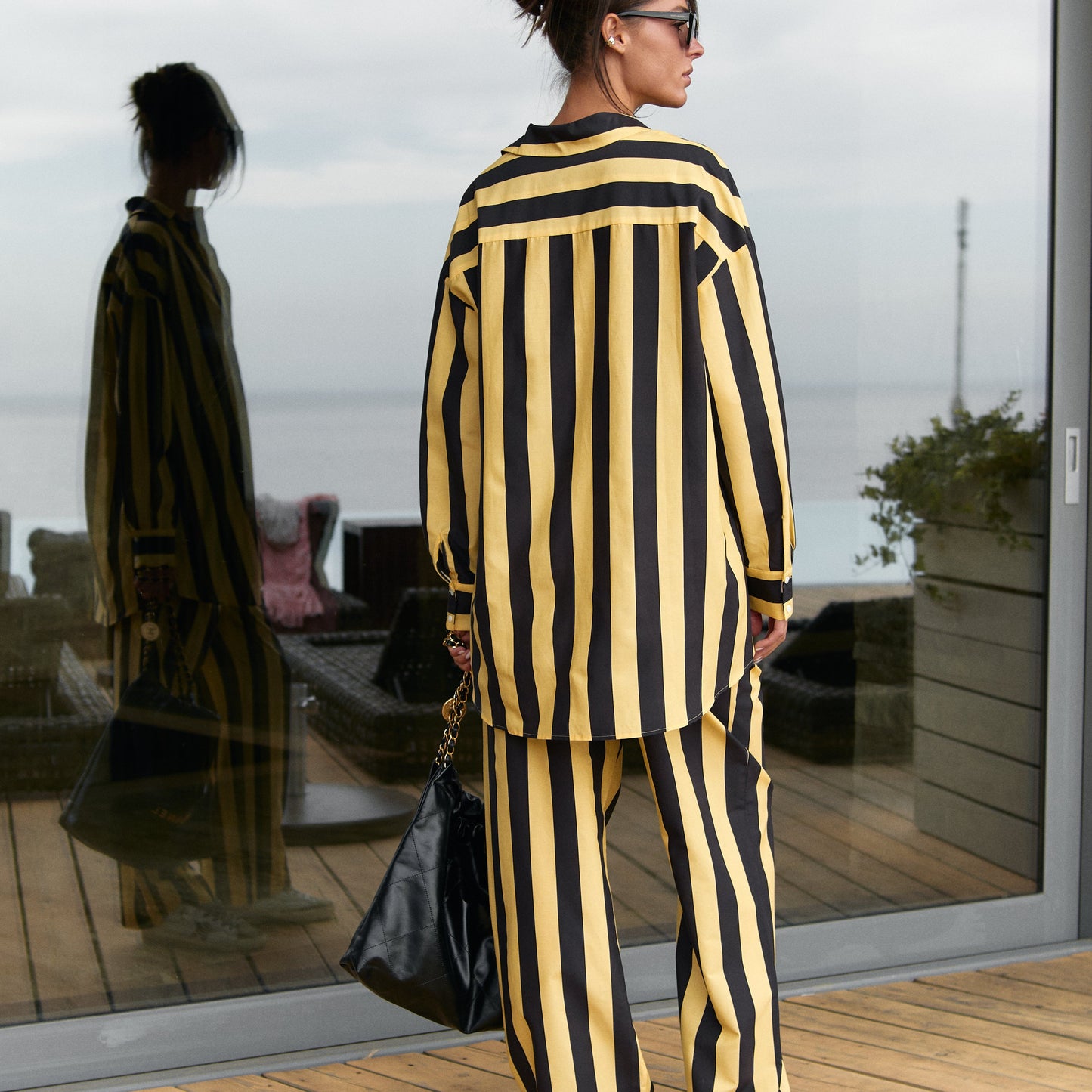 A person wearing a black and yellow striped cotton silk shirt and pants, Nokaya, is standing on a wooden deck with a reflective glass wall in the background.