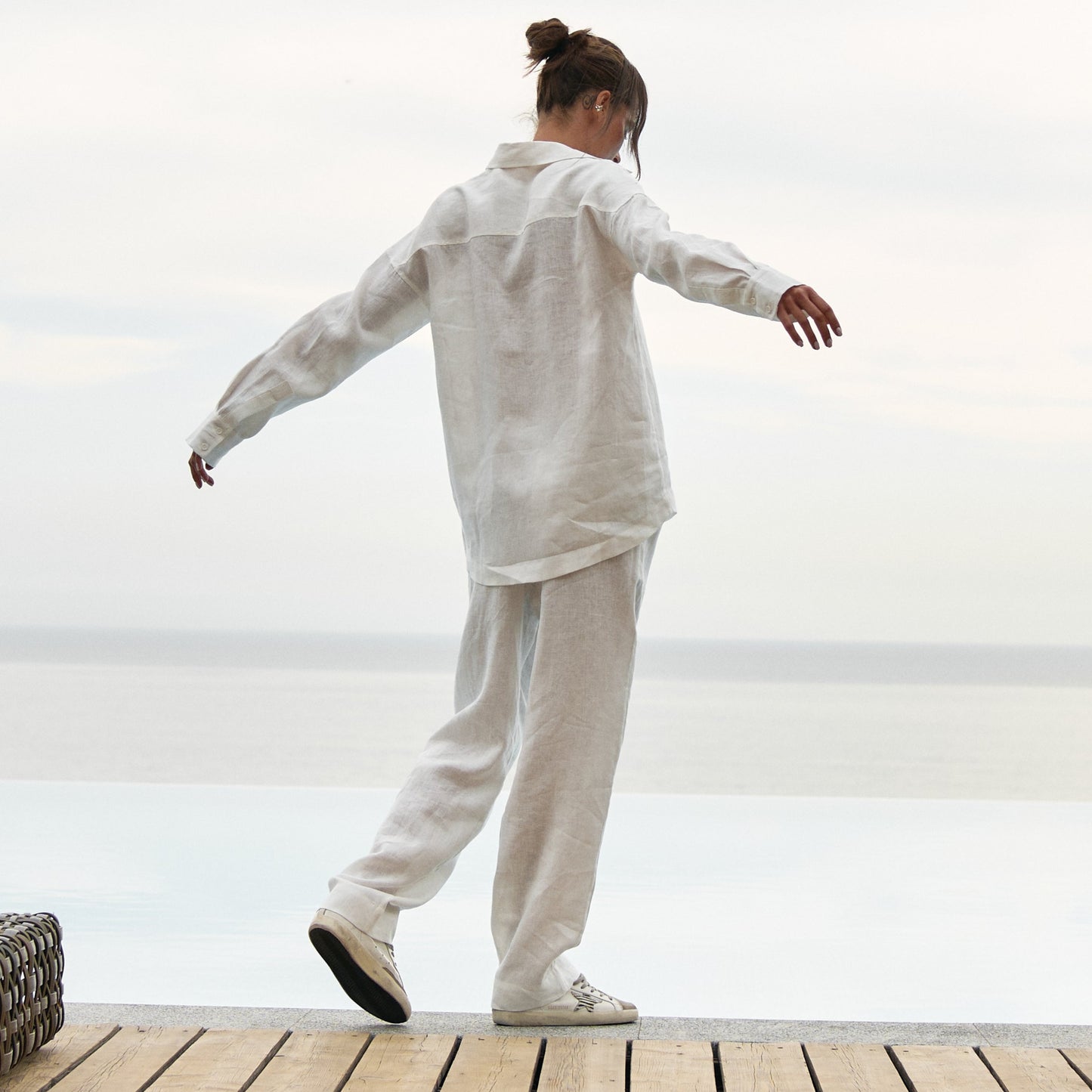 Person in white outfit standing on a wooden deck with a white wall and chair in the background