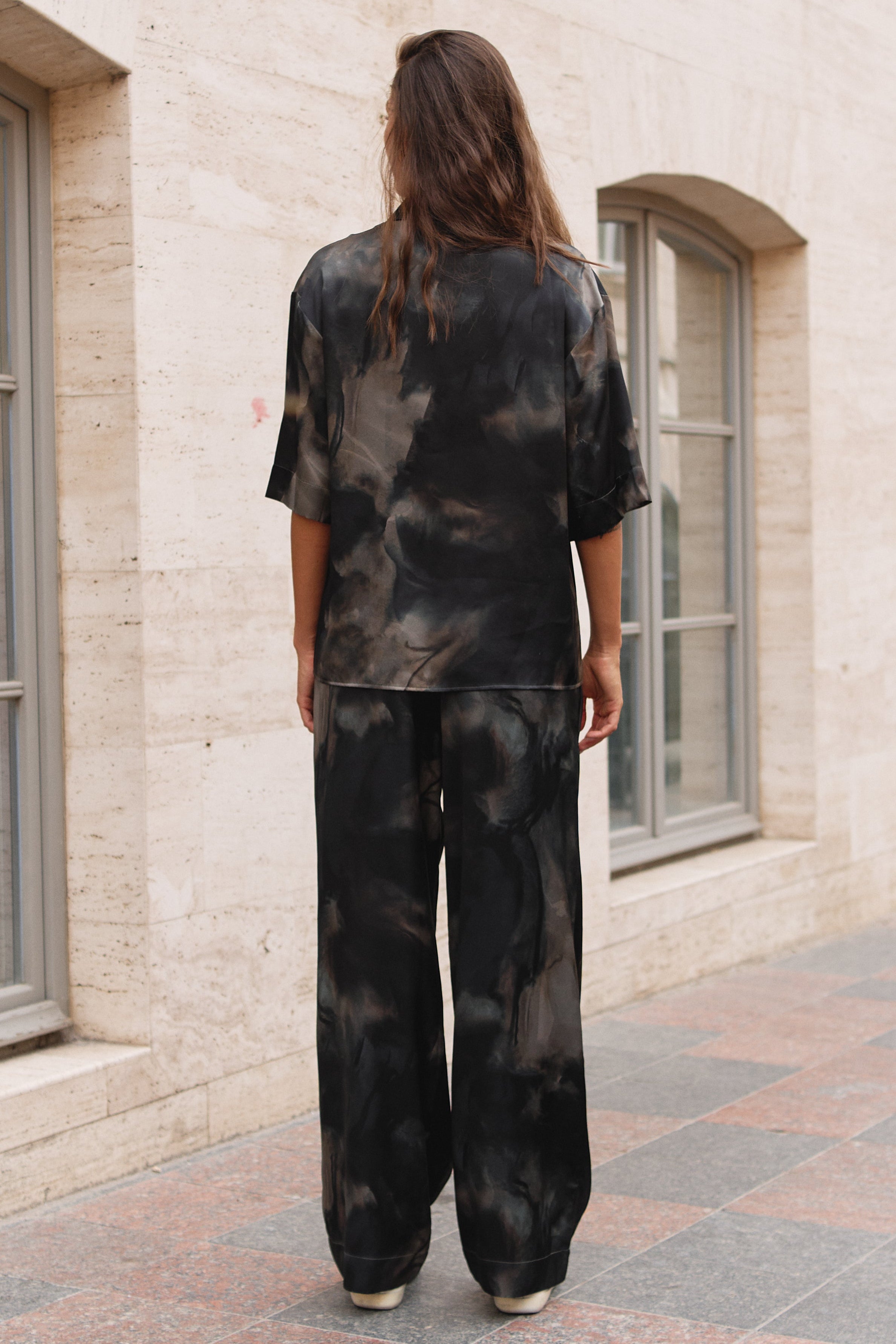 A woman viewed from behind wearing a matching black and gray tie-dye lounge set consisting of a short-sleeve top and wide-leg pants, standing on a paved walkway in front of a beige stone building.