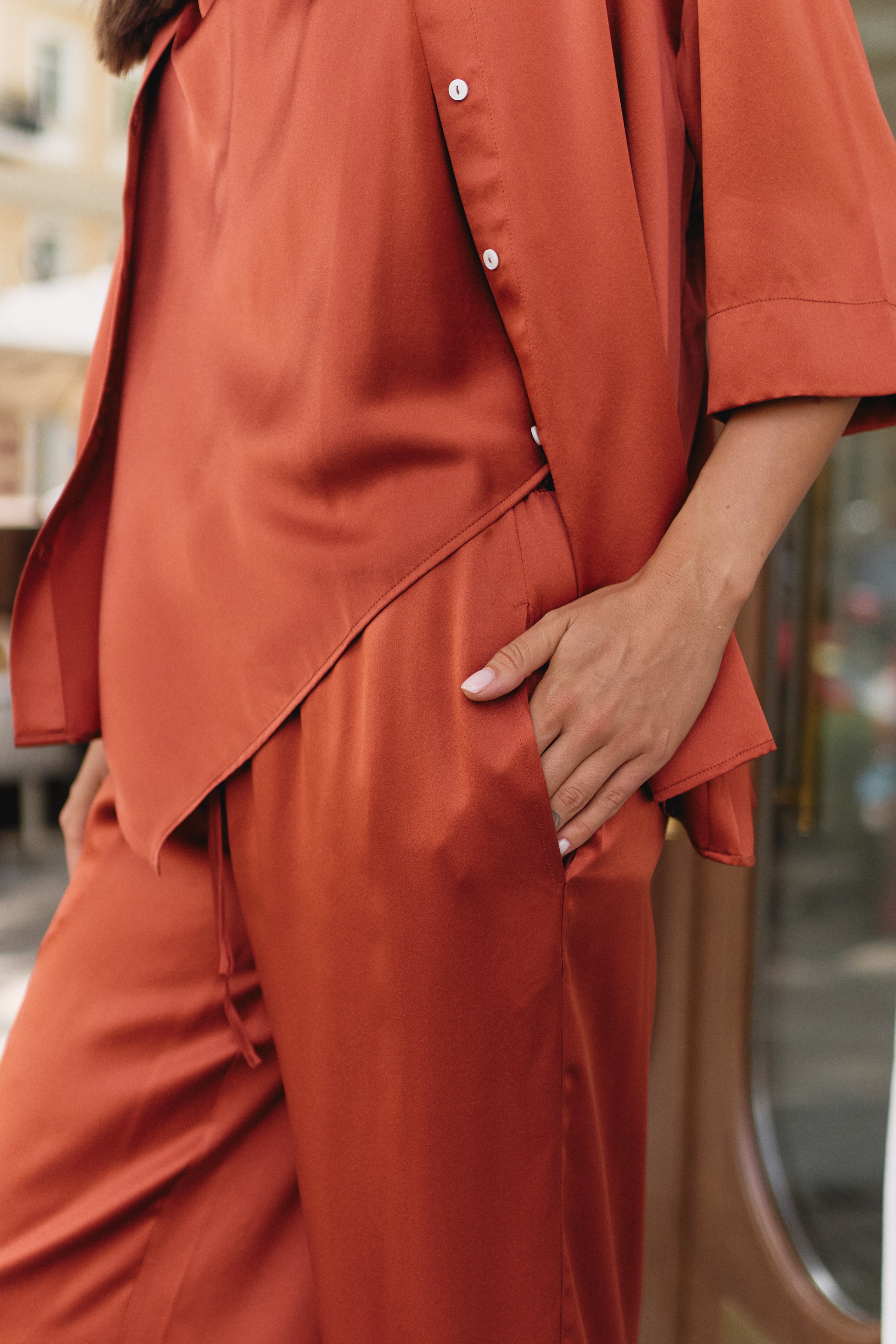 A close-up of a person wearing a matching rust-orange satin pajama set with a button-front top and drawstring pants, photographed outdoors with a softly blurred street background.
