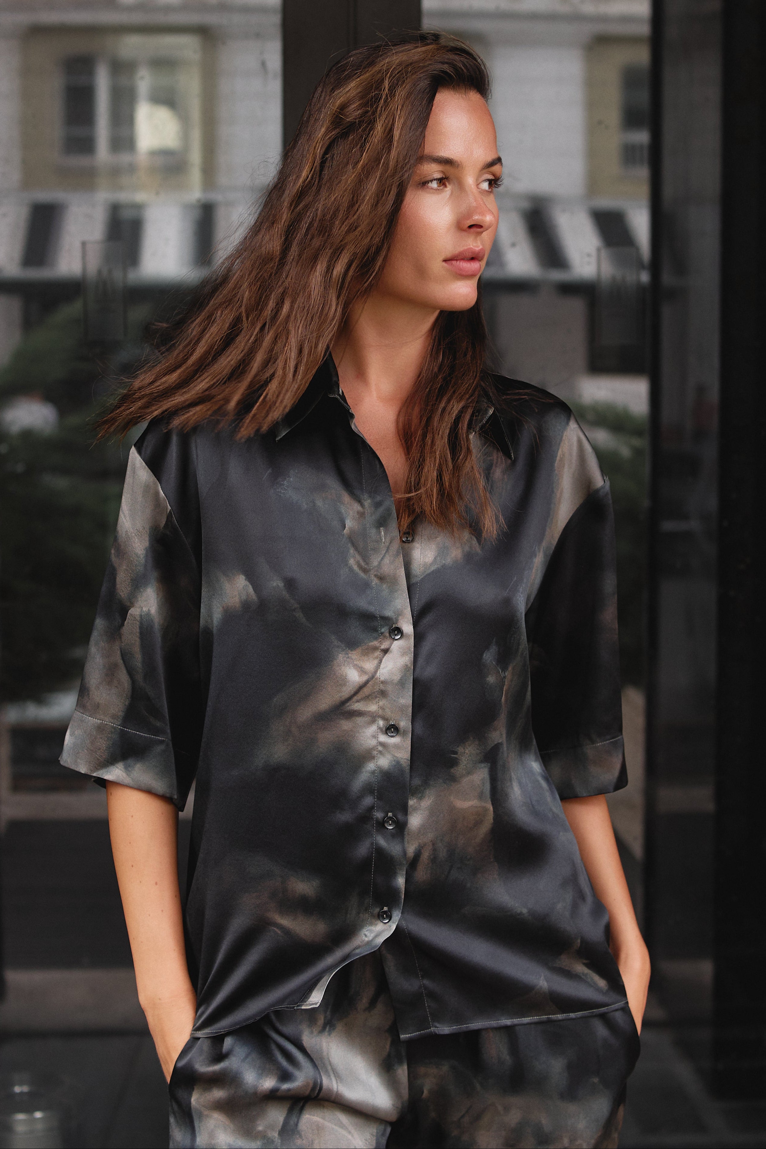  black and grey silk satin set with a tie-dye pattern Nokaya, standing in front of a glass door with a blurred city background.