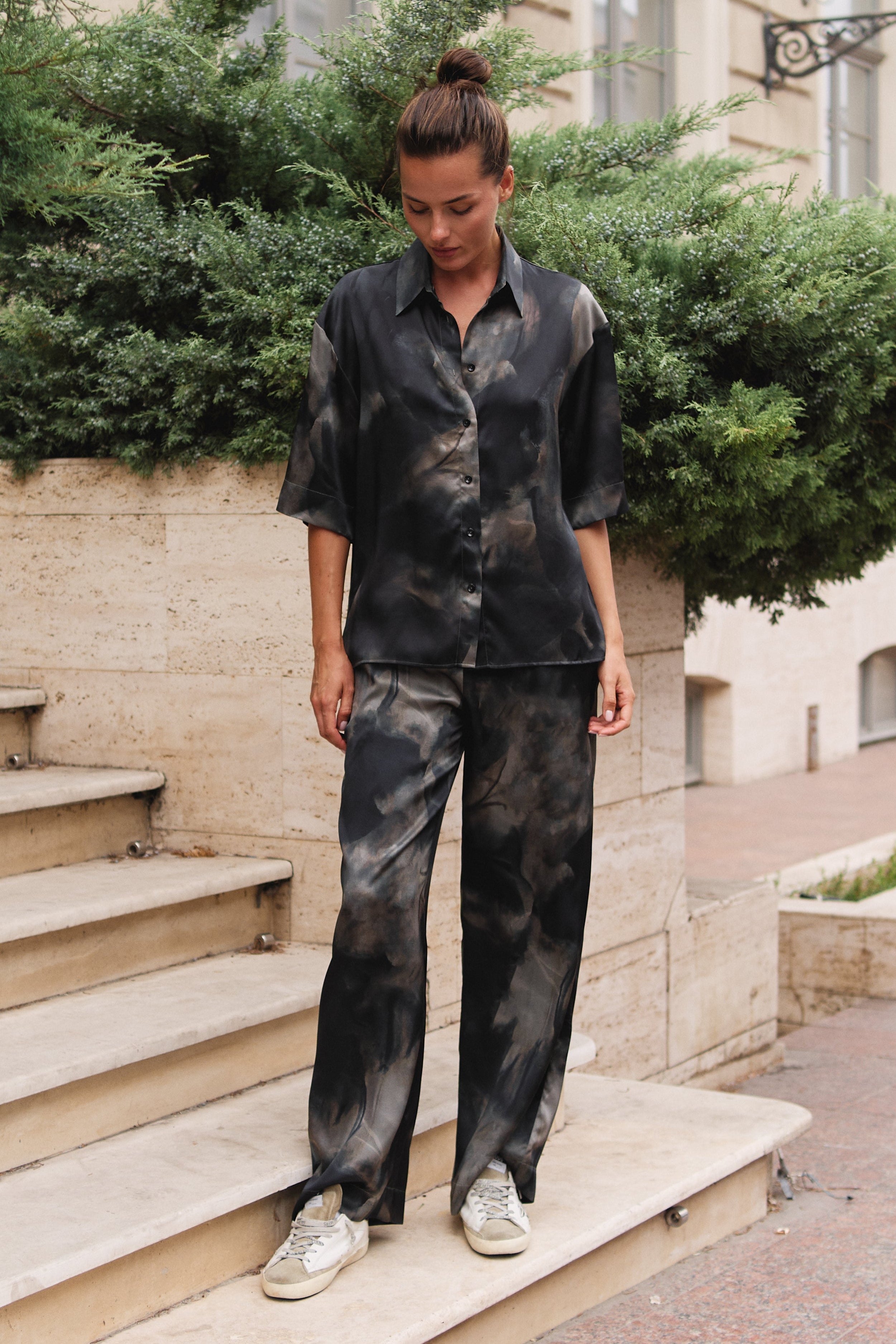  Matching black and grey tie-dye button-up silk shirt and wide-leg pants, Nokaya stands on outdoor stone steps in front of a beige building with greenery.