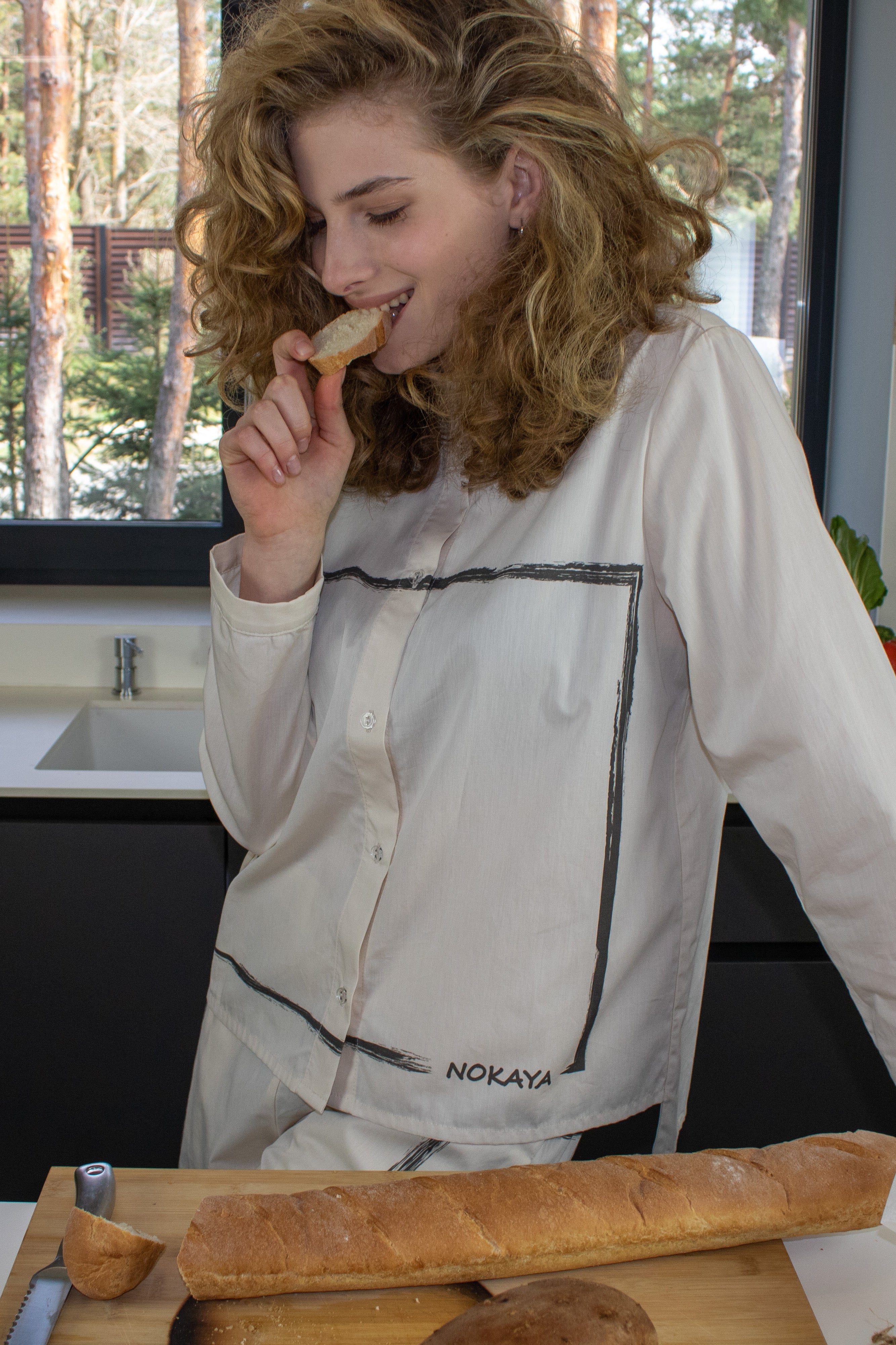 A woman in a cotton shirt, Nokaya, in a kitchen, eating bread with a view of trees outside the window.