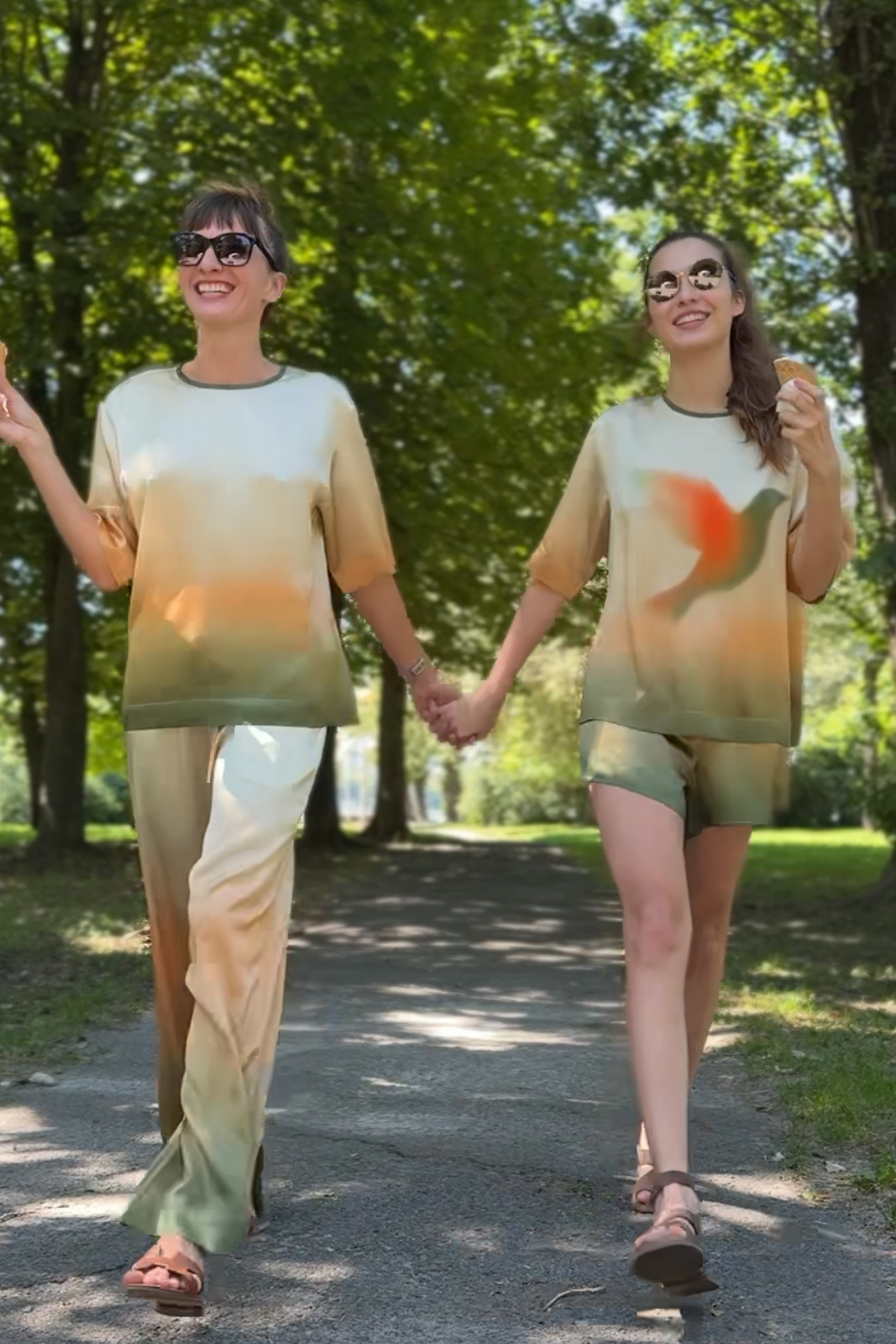 Two women walking hand-in-hand on a tree-lined path, wearing matching tie-dye lounge sets in beige, yellow, and orange. The image has black letterbox bars at the top and bottom.