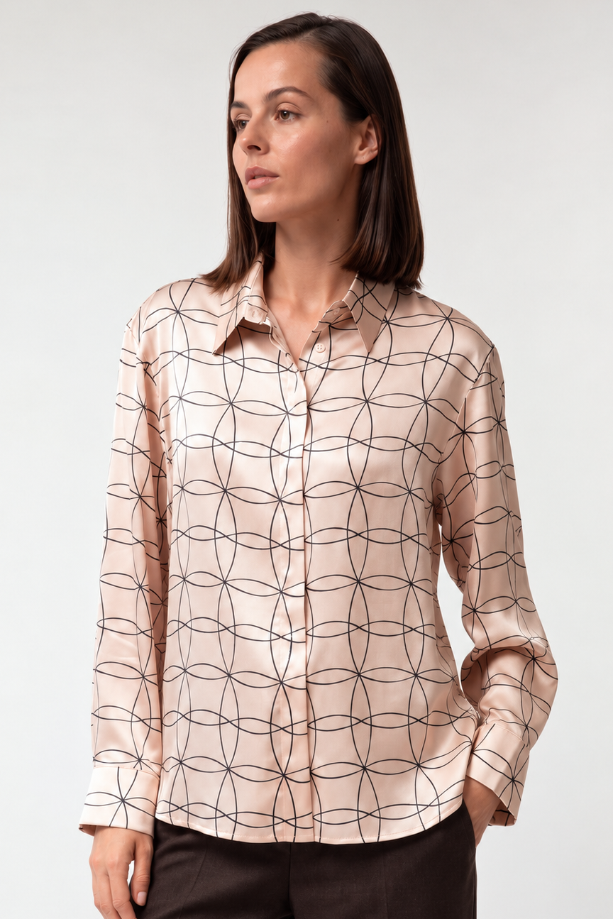 A woman modeling a long-sleeve button-up shirt with a beige base and a black geometric interlocking circle pattern, paired with dark brown trousers, against a light gray studio background.