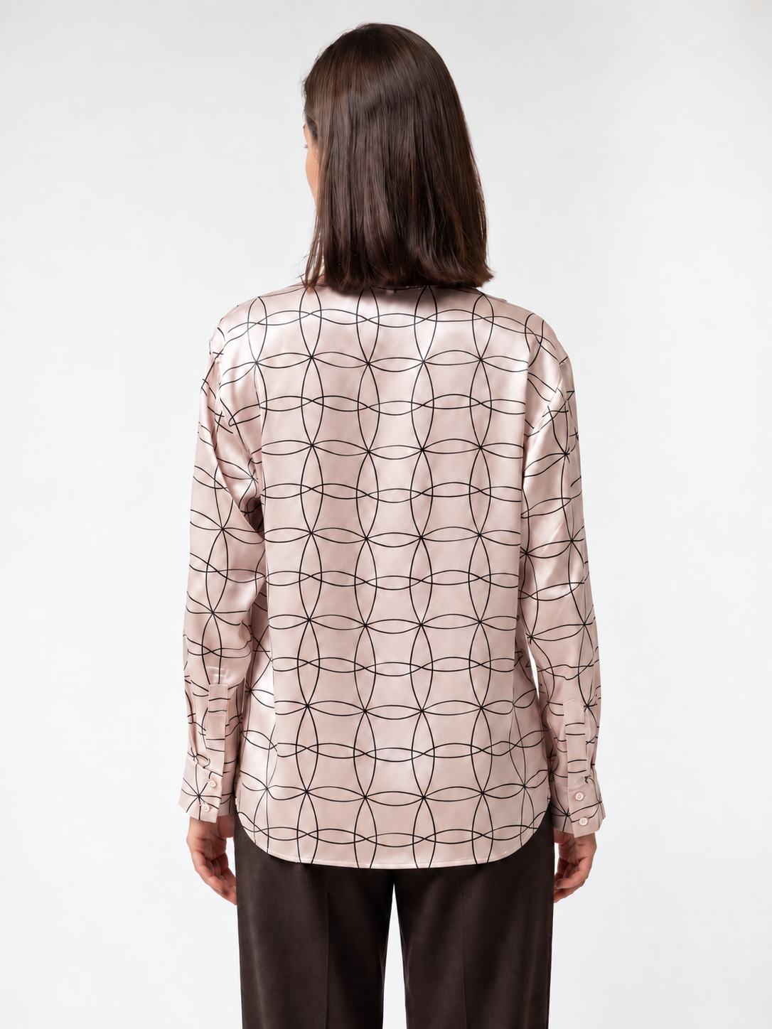 A woman viewed from the back wearing a long-sleeve satin blouse with a black geometric interlocking circle pattern on a light pinkish-beige background, paired with dark brown trousers, against a plain white studio background.