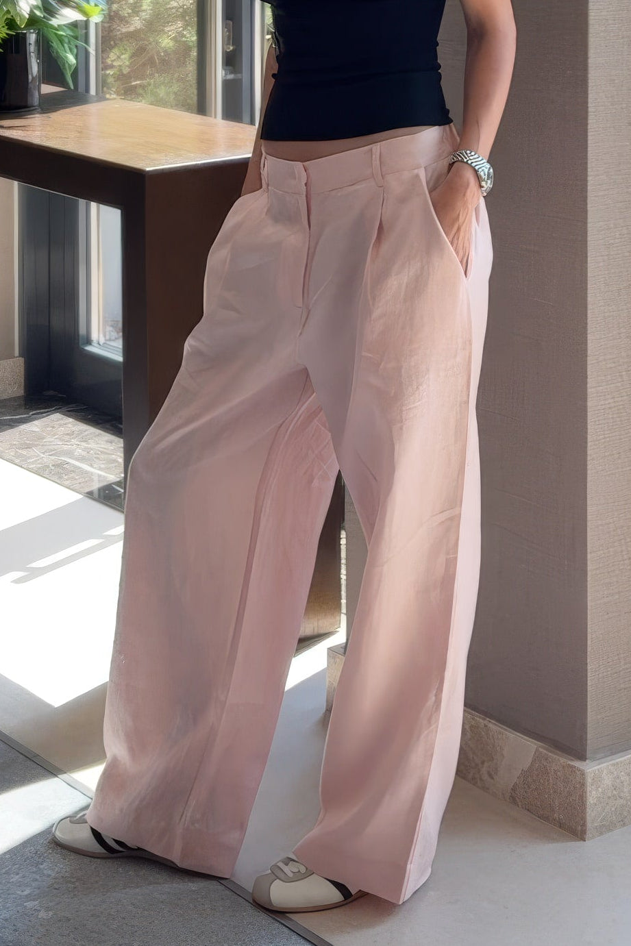 Linen pink wide-leg trousers Nokaya stand in a modern indoor space with large windows, neutral walls, and a console table with flowers.