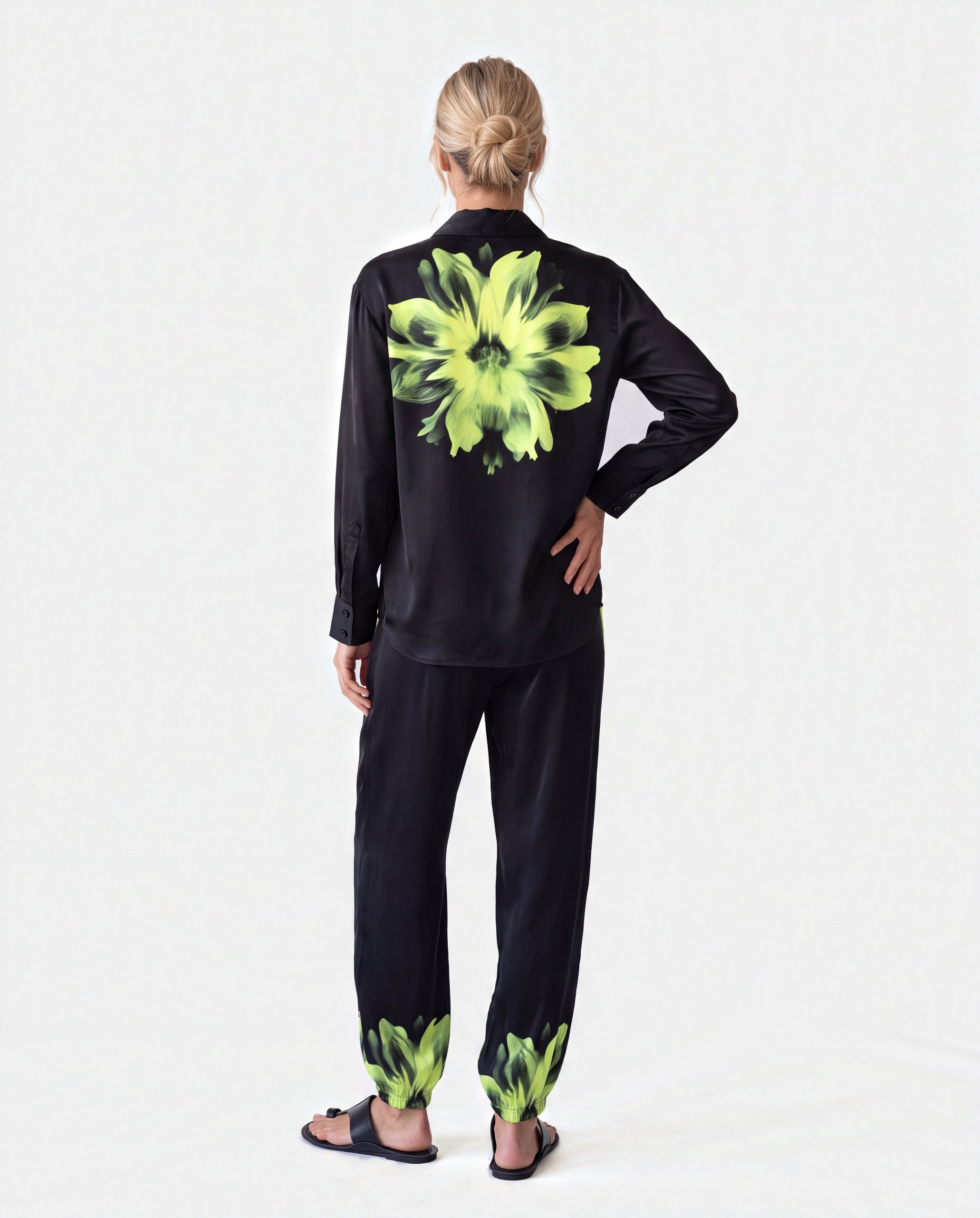 A woman viewed from the back wearing a black long-sleeve shirt and matching pants with a large neon yellow-green floral print on the back and matching print near the cuffs, photographed against a plain light gray studio background. Nokaya