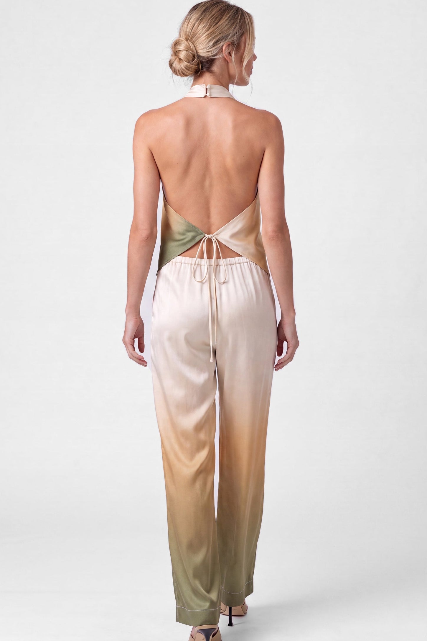 A woman shown from the back wearing a silk satin halter top and pants, Nokaya, with a gradient colour transition from olive green to beige, paired with black high heels, against a light grey studio background.