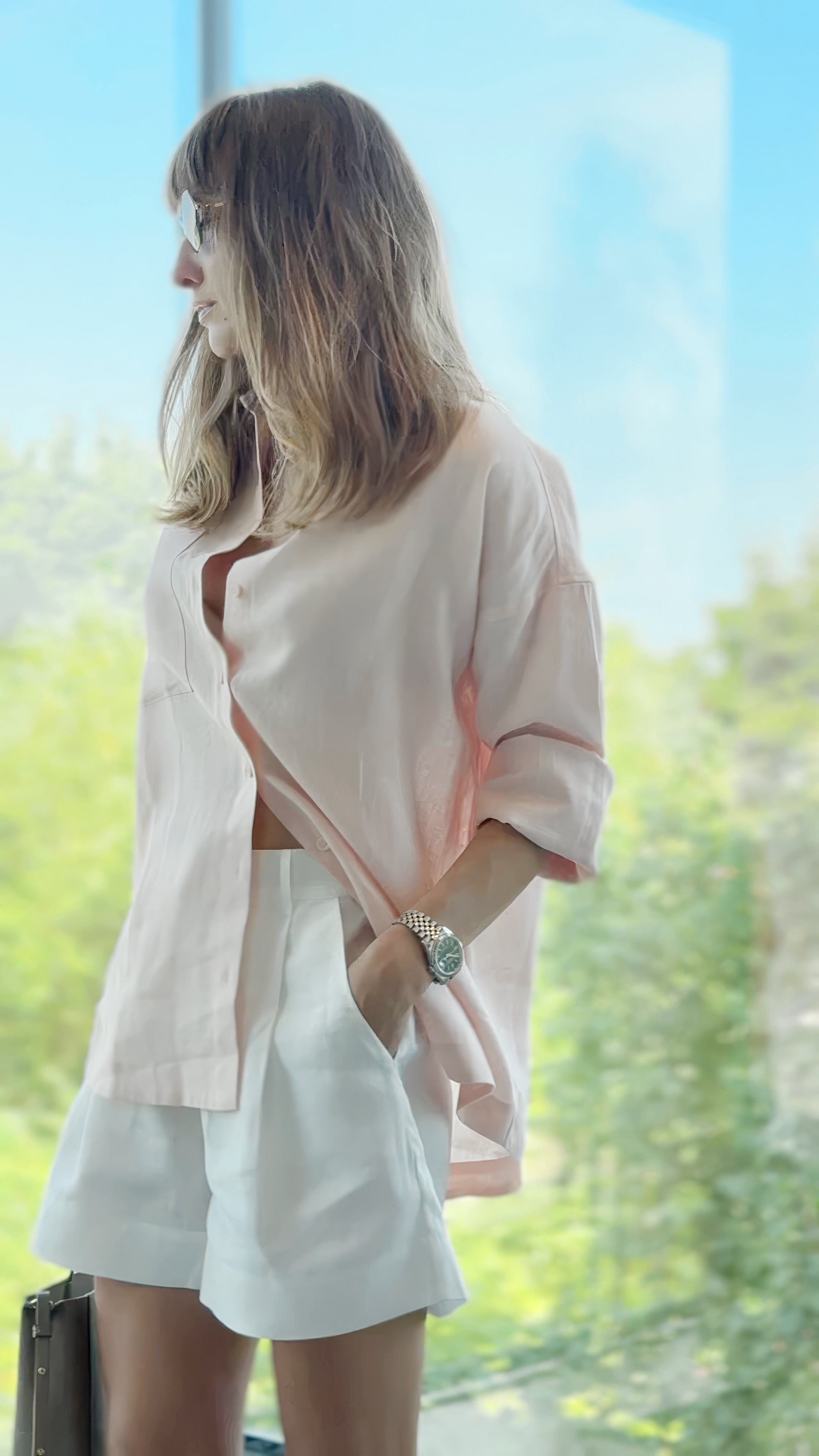 A woman wearing a light pink oversized button-up shirt and white linen shorts Nokay, standing in front of a large window with a green outdoor view.