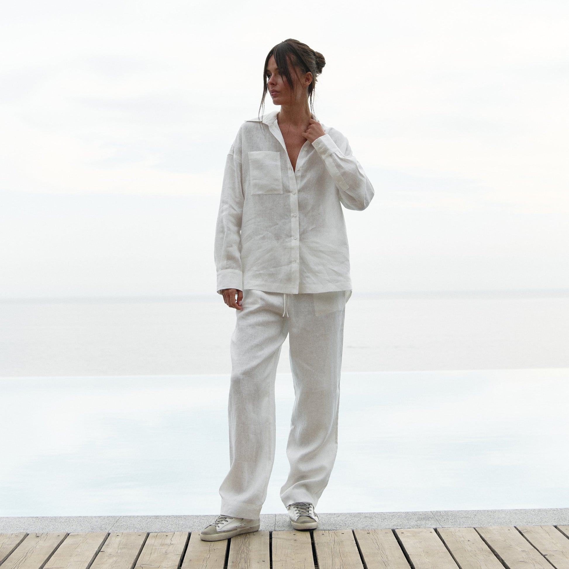 A woman wearing a white linen outfit is standing on a wooden deck with a white background. Linen Spirit collection by Nokaya.