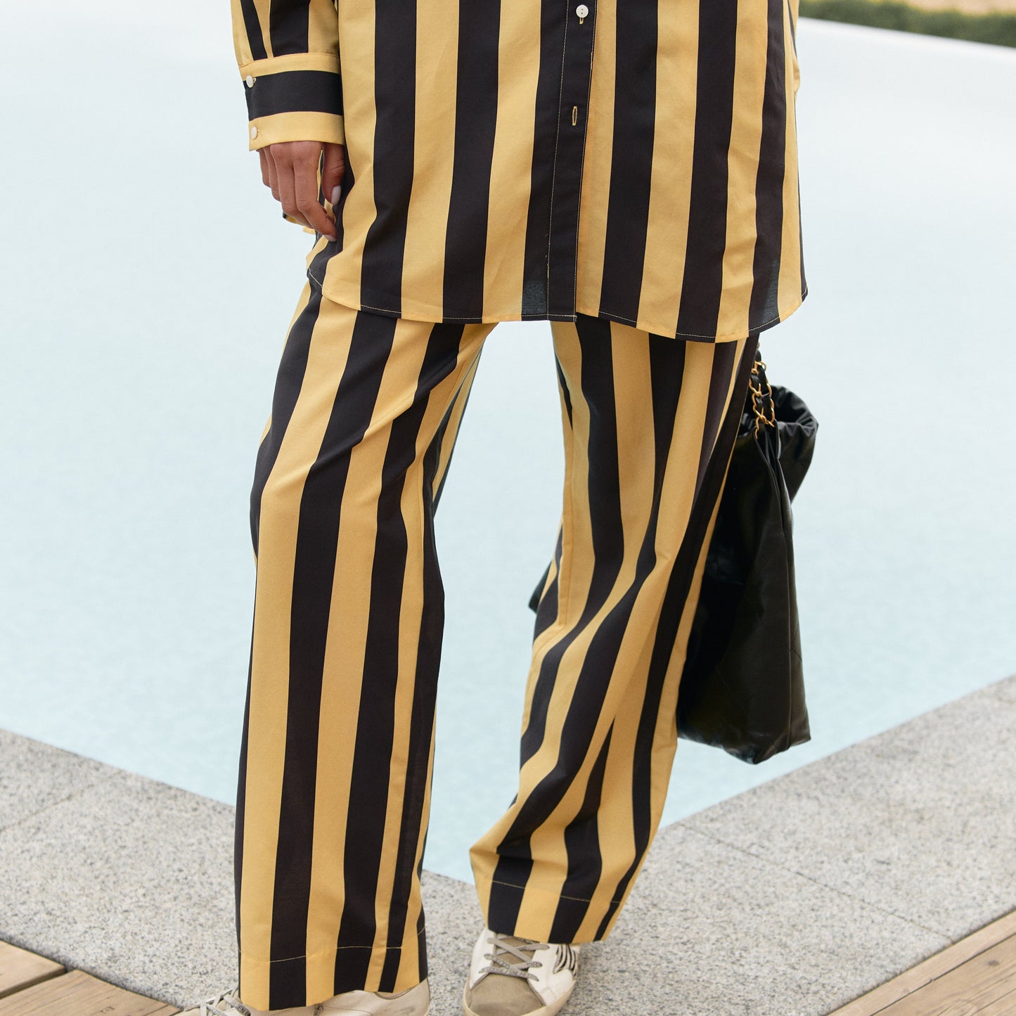 A person wearing black and yellow striped cotton pants is standing on a wooden deck by a body of water. Nokaya summer 2025