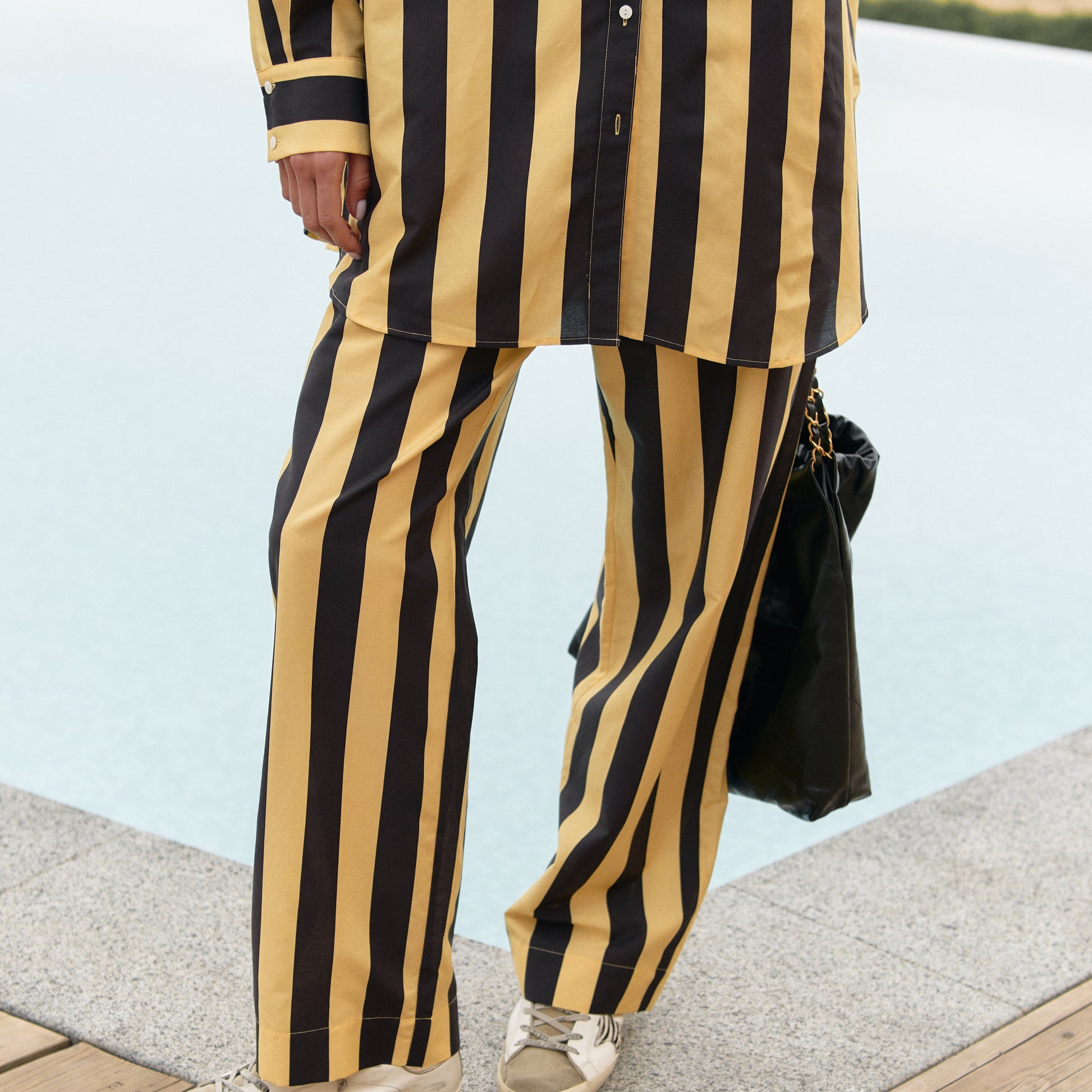 A person wearing black and yellow striped cotton pants is standing on a wooden deck by a body of water. Nokaya summer 2025
